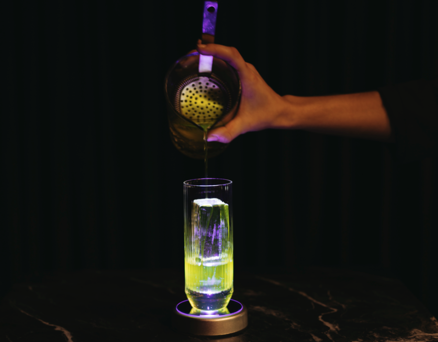 Eine Hand füllt in ein Highball Glas ein grünlichen Cocktail mit einem großen rechteckigen Eiswürfel. Das Glas wird durch den Coaster von unten beleuchtet und wirkt somit noch heller und cooler
