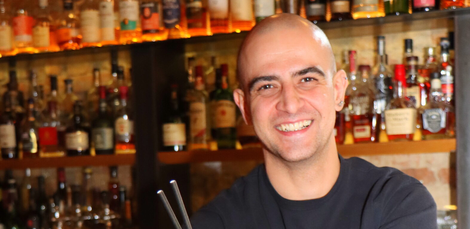 Der Barkeeper steht hinter dem Bartresen und hält einen gelben Cocktail in der Hand. Dabei schaut er direkt in die Kamera und lächelt.