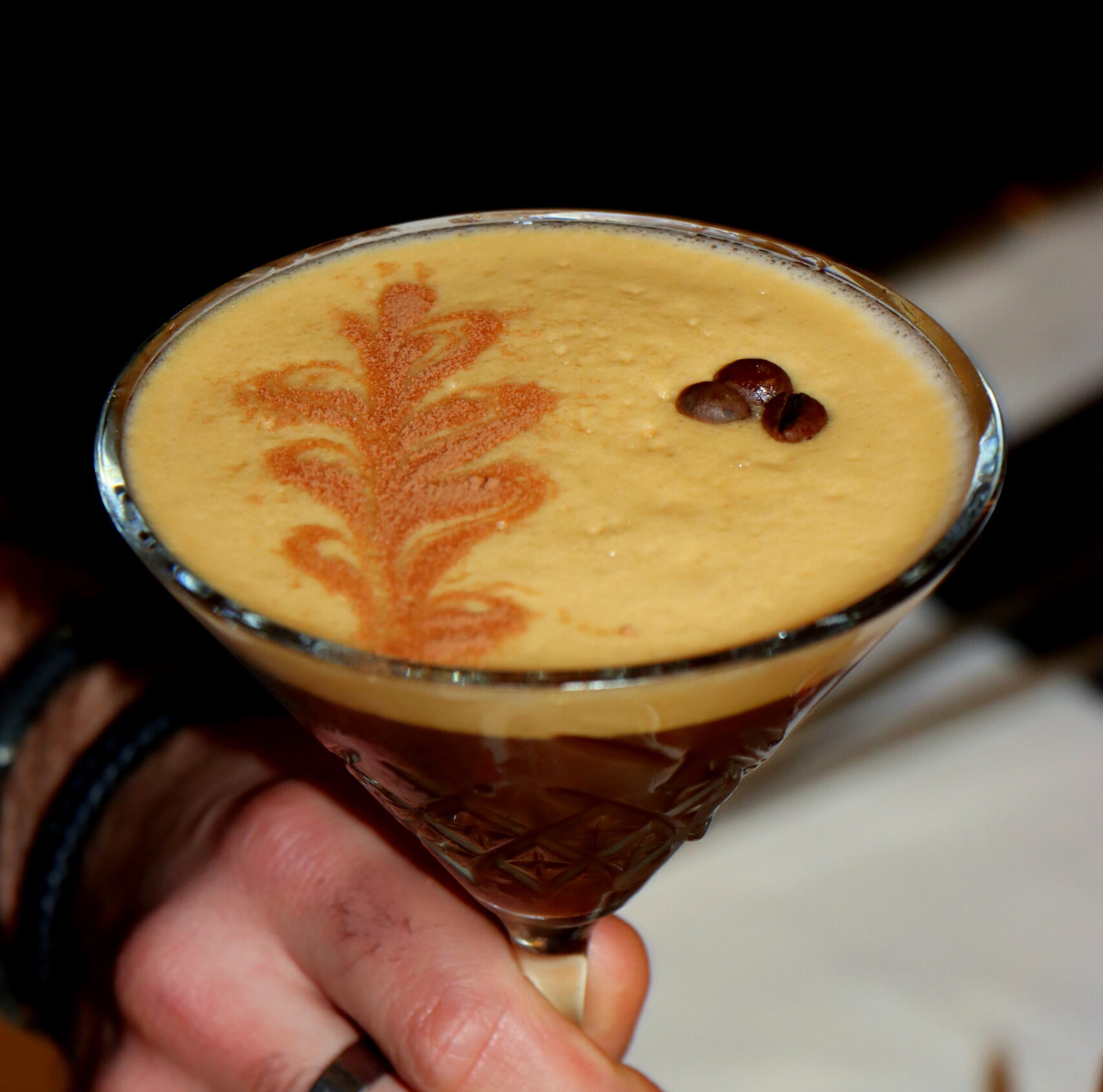 Eine Hand hält einen Espresso Martini. Der Schaum ist sehr schön und mit einem aus Kaffeegezeichneten Blatt verziert.