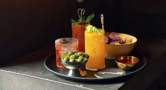 Auf einem Tisch steht ein Tableau mit 3 verschieden farbigen Cocktails, vorne sind Oliven in einer Schale und im Hintergrund steht eine Kerze.